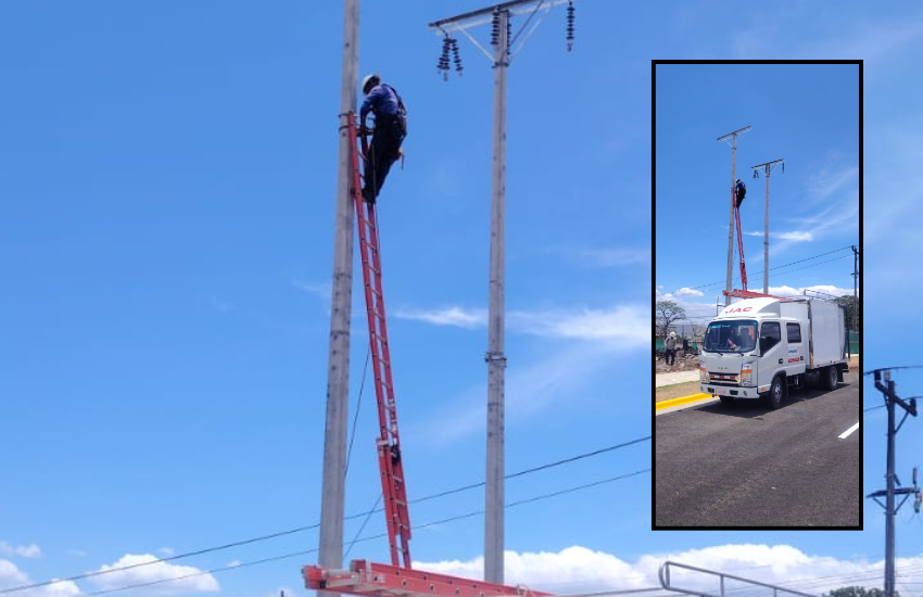 Un trabajador con equipo de seguridad trepando a un poste alto de servicios públicos junto a una carretera, con un camión utilitario blanco estacionado debajo y un cielo azul de fondo.